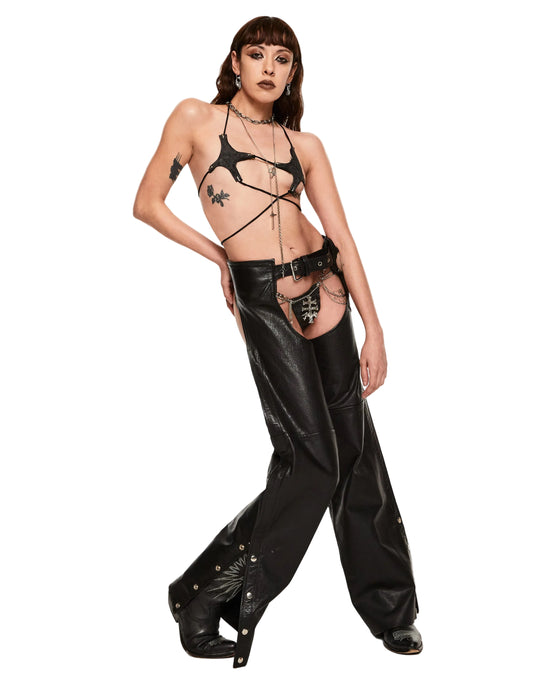 A model poses in a studio setting wearing a black strappy harness-style bra top and matching wide-leg black faux-leather pants with gold-tone hardware.