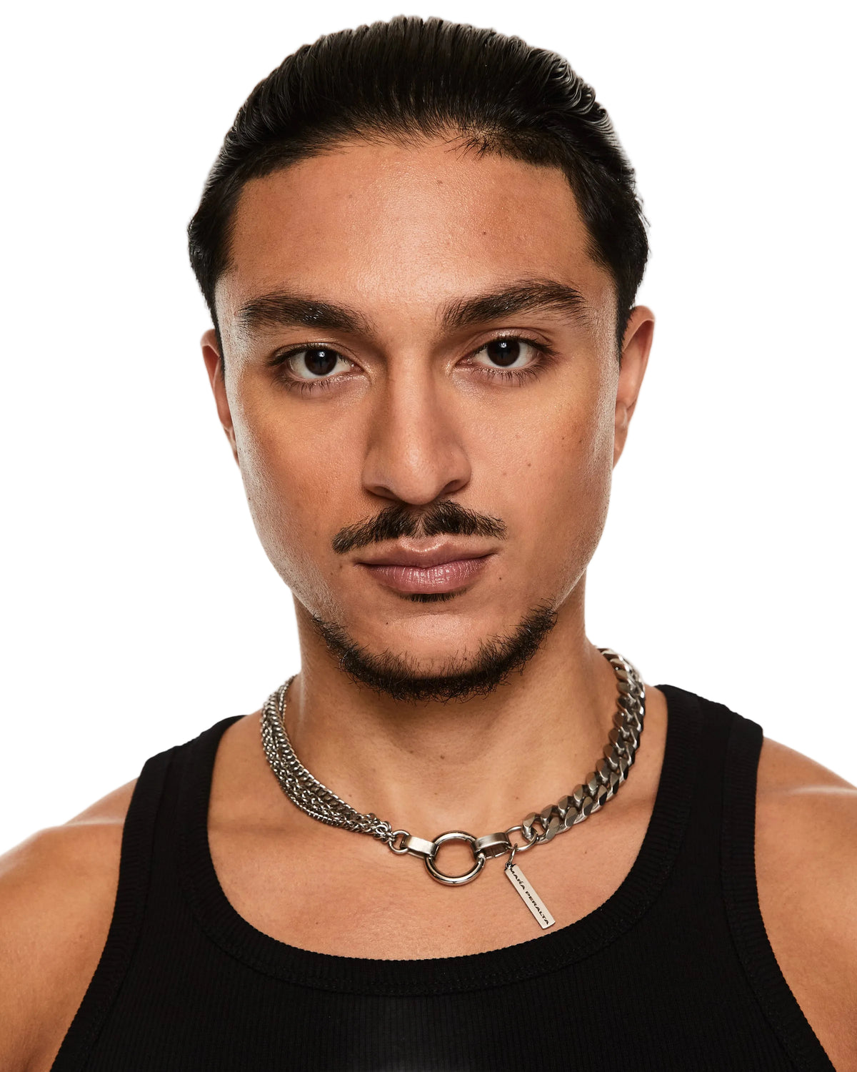 A close-up portrait of a man with slicked-back dark hair wearing a chunky silver chain necklace with a ring clasp and a black ribbed tank top against a plain white background.