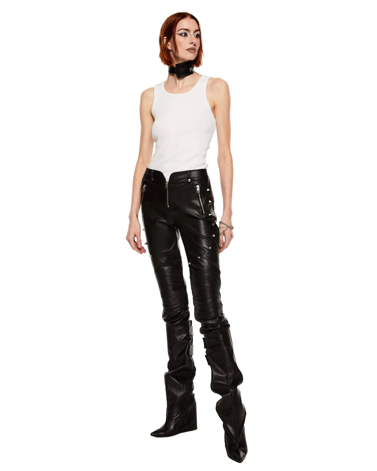 A model stands against a plain white studio background wearing a white sleeveless top, black leather pants with multiple zippers and studs, and tall black pointed boots.