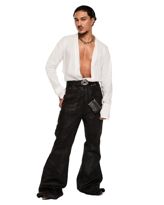 A model stands against a white studio background wearing a white long-sleeve button-up shirt tied loosely at the waist, black wide-leg jeans with a silver belt and silver buckle, layered necklaces, and black shoes.
