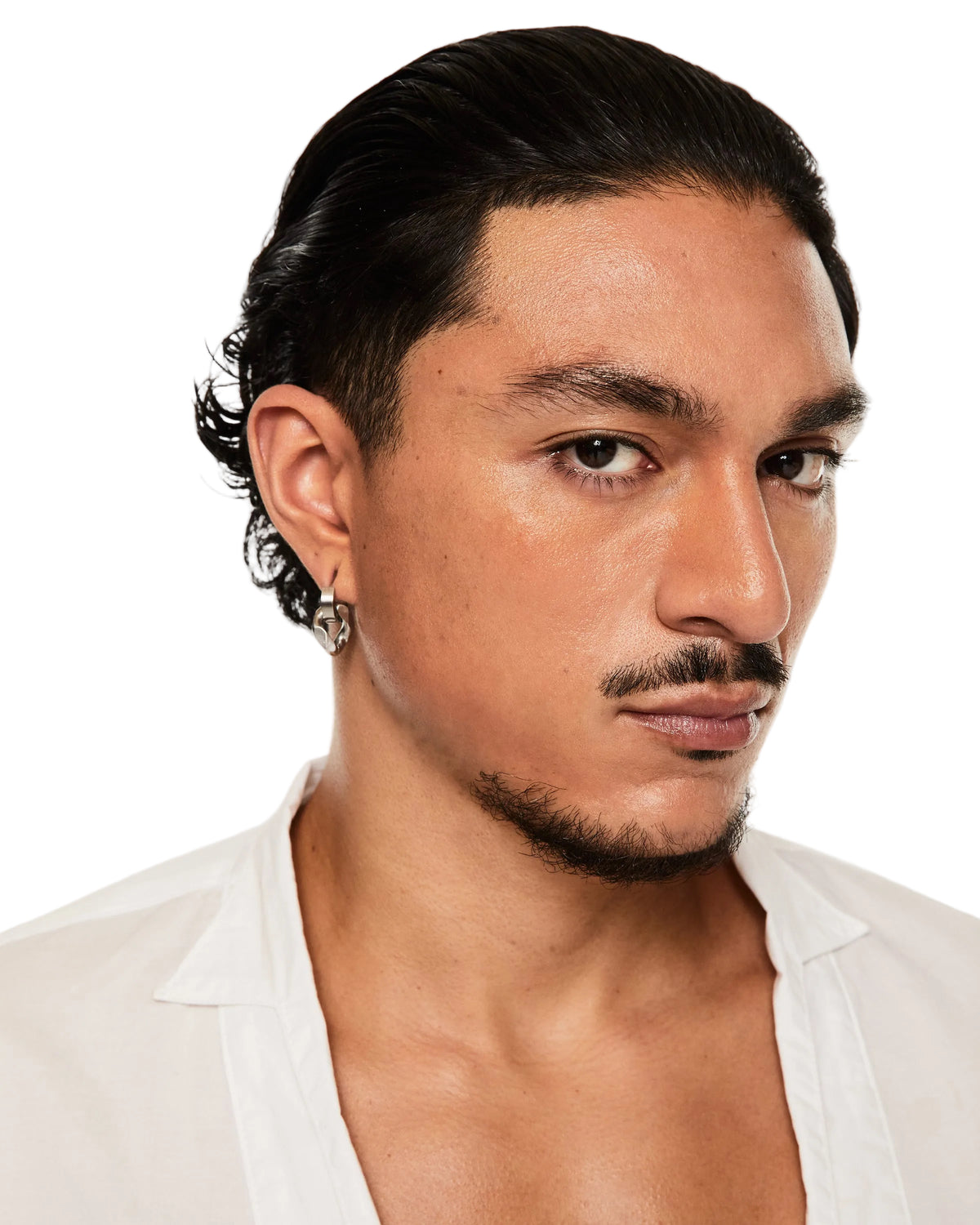 A close-up portrait of a man with slicked-back dark hair, wearing a silver geometric hoop earring on his left ear, a white open-collar shirt, and a small mustache and goatee. The background is a clean, solid white studio backdrop.