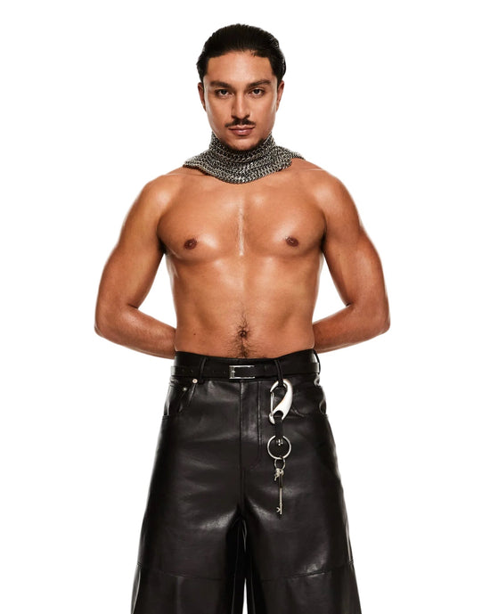 A shirtless male model wearing black wide-leg faux leather pants with silver hardware, a black belt, and a silver chain necklace, standing against a white studio background.